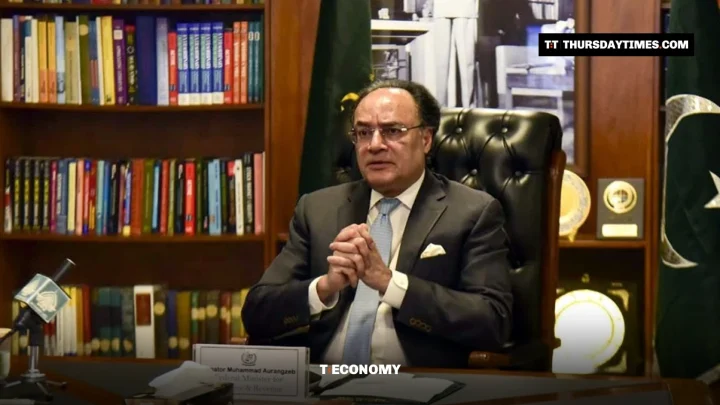 Muhammad Aurangzeb in a grey suit sits at his desk with clasped hands, Pakistani flags and bookshelves behind him